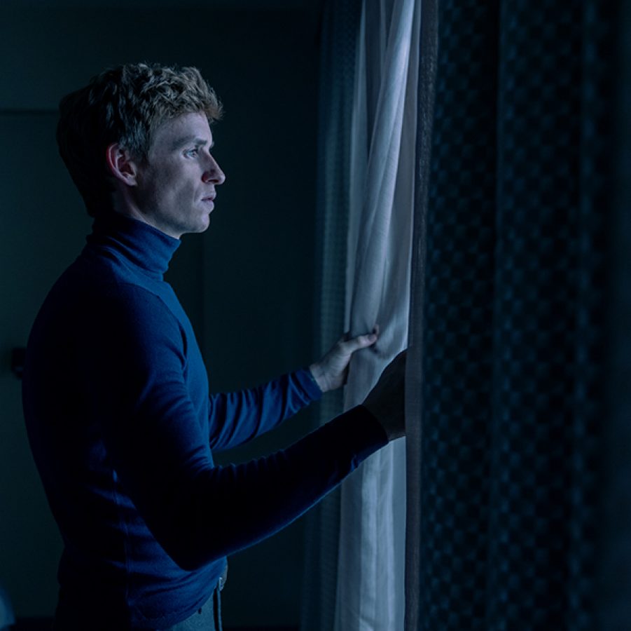 In a scene from The Day of the Jackal, a smartly dressed man (Eddie Redmayne) stands by a window in a dark room, pulling back a curtain to look outside. 